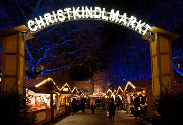 This may contain: the entrance to christmas market lit up at night with people walking under it and trees in the background