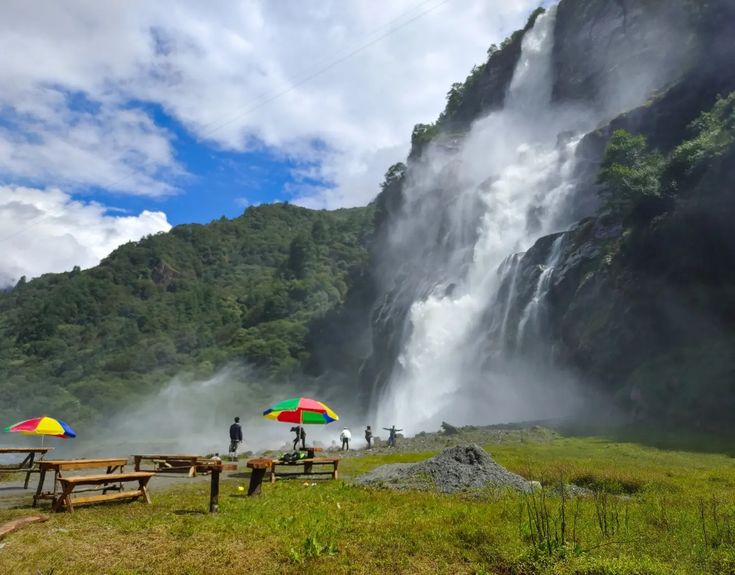 This may contain: two people with umbrellas are standing in front of a waterfall and picnic tables under them