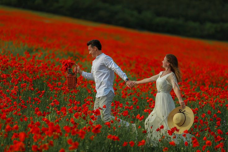 This may contain: a man and woman holding hands in a field full of red flowers with a picnic basket