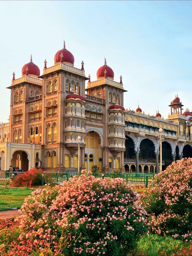 This may contain: an ornate building with red domes on top and pink flowers in the foreground, surrounded by greenery