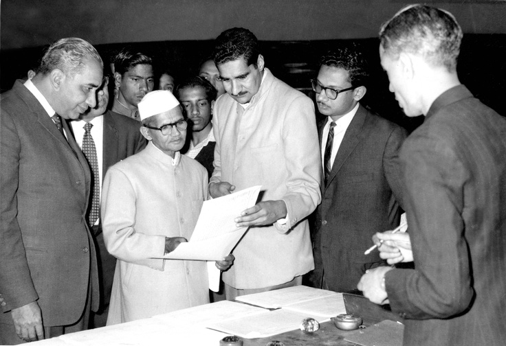 This may contain: black and white photograph of men in suits looking at papers on a table with other men