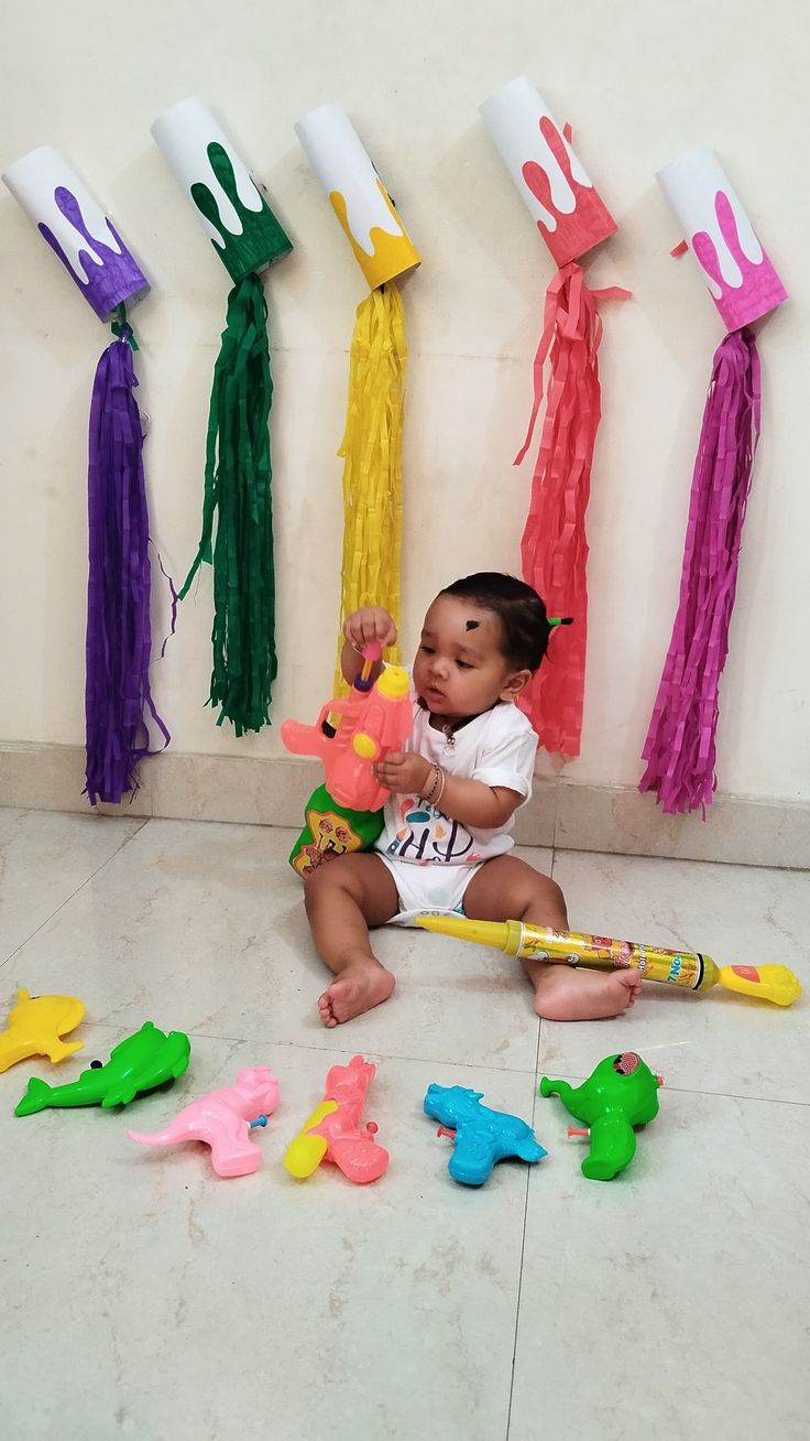 This may contain: a small child sitting on the floor with toys in front of her and several colorful streamers hanging from the wall behind him