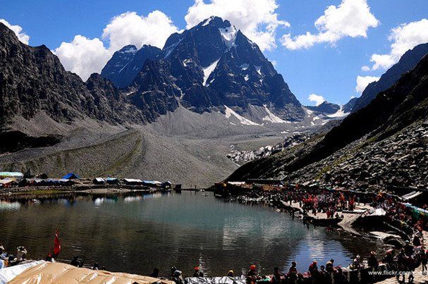 This may contain: a group of people standing around a lake with mountains in the background