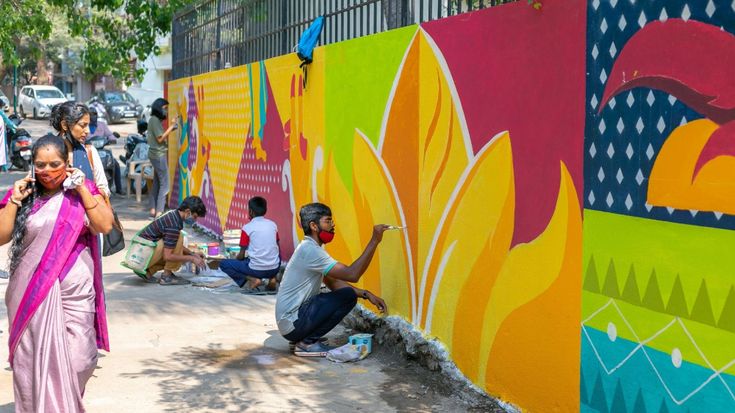 This may contain: a woman in a pink sari painting a colorful wall with people sitting on the ground