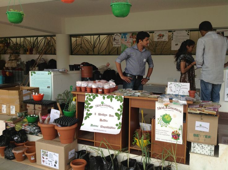 This may contain: two men are standing behind a table with plants and seed packets on it in front of other people