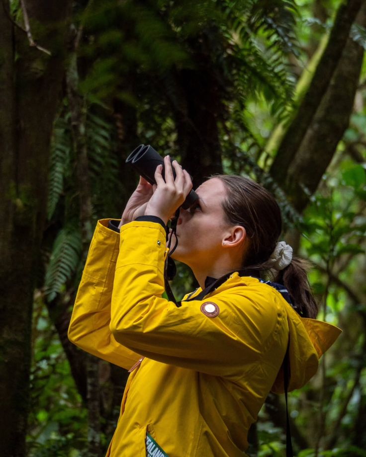 This may contain: a woman wearing a yellow jacket is looking into the sky with binoculars in her hand